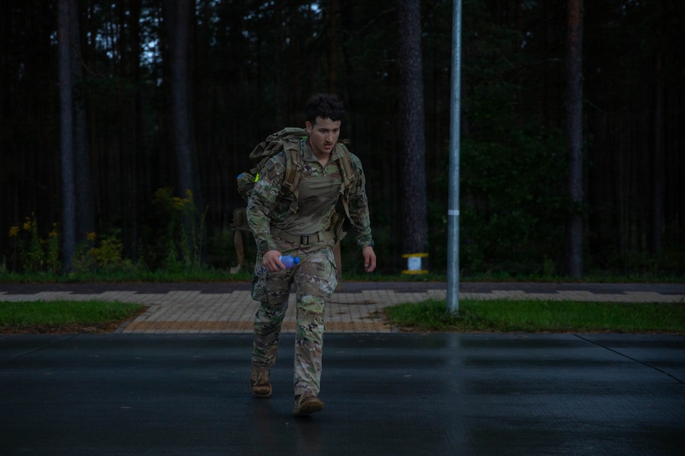 A soldier in camouflage uniform and full tactical gear is moving at pace along a wet road in low-light conditions, appearing physically exerted. They are carrying a loaded rucksack on their back and holding a water bottle in one hand. The setting appears to be a military installation or training area bordered by a dense forest.