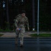 A soldier in camouflage uniform and full tactical gear is moving at pace along a wet road in low-light conditions, appearing physically exerted. They are carrying a loaded rucksack on their back and holding a water bottle in one hand. The setting appears to be a military installation or training area bordered by a dense forest.