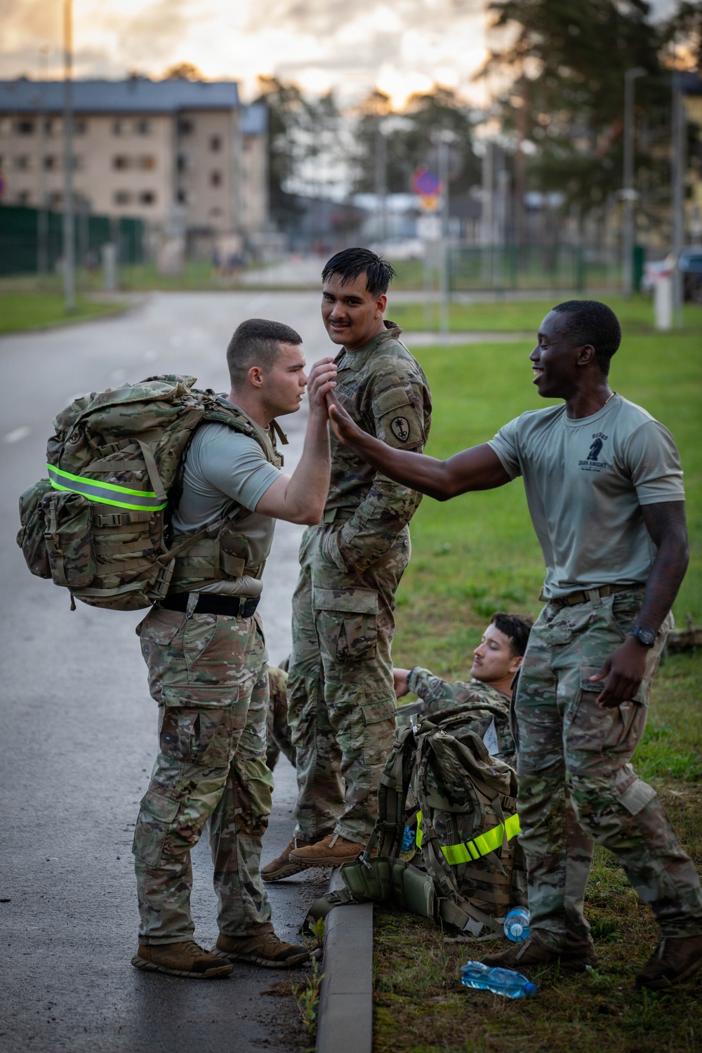 Four military personnel in U.S. Army OCP (Operational Camouflage Pattern) uniforms are gathered near a road at dusk or dawn, appearing to be at the conclusion of a ruck march or physical training event. Two soldiers are standing and interacting in a congratulatory or supportive manner, one soldier is seated on the ground resting, and one soldier is wearing a t-shirt without a jacket. The setting appears to be a military installation based on the background buildings and infrastructure.