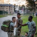 Four military personnel in U.S. Army OCP (Operational Camouflage Pattern) uniforms are gathered near a road at dusk or dawn, appearing to be at the conclusion of a ruck march or physical training event. Two soldiers are standing and interacting in a congratulatory or supportive manner, one soldier is seated on the ground resting, and one soldier is wearing a t-shirt without a jacket. The setting appears to be a military installation based on the background buildings and infrastructure.
