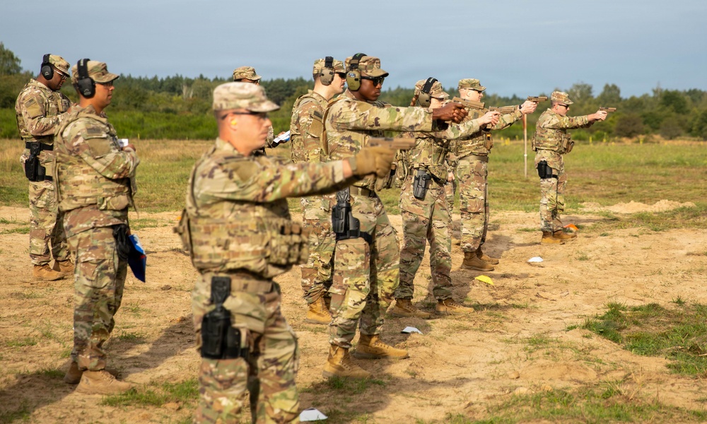 A group of military personnel in MultiCam/OCP camouflage uniforms are conducting pistol qualification or training on an outdoor range. Several soldiers are in shooting stances with handguns extended downrange, while at least one individual appears to be observing or instructing. The setting is an open sandy field with treeline in the background under overcast skies.
