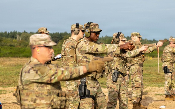 A group of military personnel in MultiCam/OCP camouflage uniforms are conducting pistol qualification or training on an outdoor range. Several soldiers are in shooting stances with handguns extended downrange, while at least one individual appears to be observing or instructing. The setting is an open sandy field with treeline in the background under overcast skies.