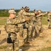 A group of military personnel in MultiCam/OCP camouflage uniforms are conducting pistol qualification or training on an outdoor range. Several soldiers are in shooting stances with handguns extended downrange, while at least one individual appears to be observing or instructing. The setting is an open sandy field with treeline in the background under overcast skies.