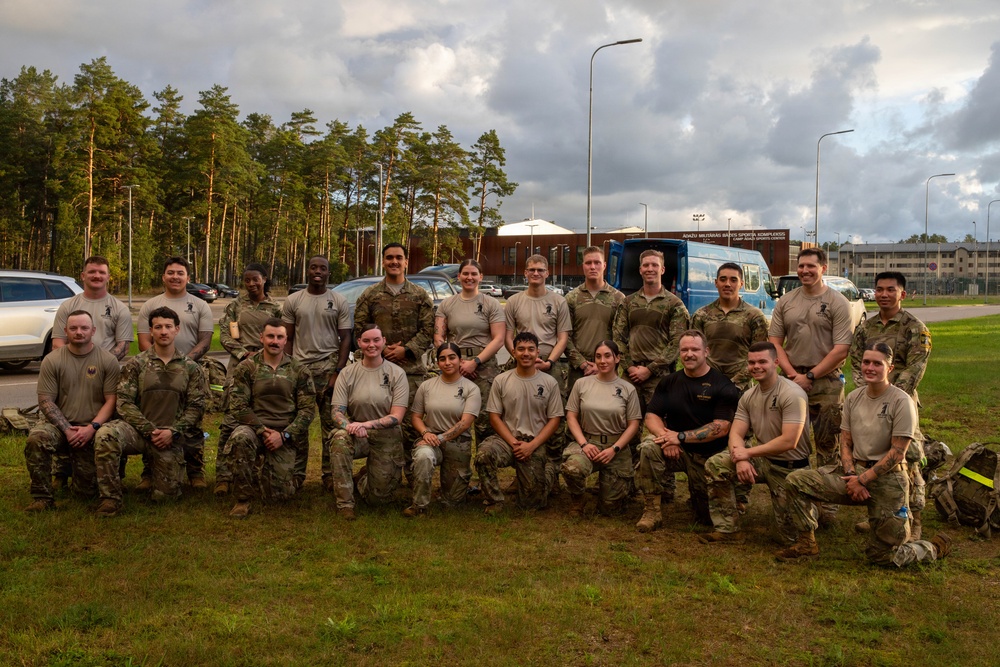 A group of approximately 22 military personnel in U.S. Army combat uniforms (OCP pattern) pose for a formal group photo on a grass area at what appears to be a military installation. The sign visible in the background reads 'Ādaži Militārās Bāzes Sporta Komplekss' (Ādaži Military Base Sports Complex), indicating this is Camp Ādaži in Latvia. The group includes both male and female soldiers arranged in two rows, with the front row kneeling and the back row standing.