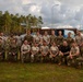 A group of approximately 22 military personnel in U.S. Army combat uniforms (OCP pattern) pose for a formal group photo on a grass area at what appears to be a military installation. The sign visible in the background reads 'Ādaži Militārās Bāzes Sporta Komplekss' (Ādaži Military Base Sports Complex), indicating this is Camp Ādaži in Latvia. The group includes both male and female soldiers arranged in two rows, with the front row kneeling and the back row standing.