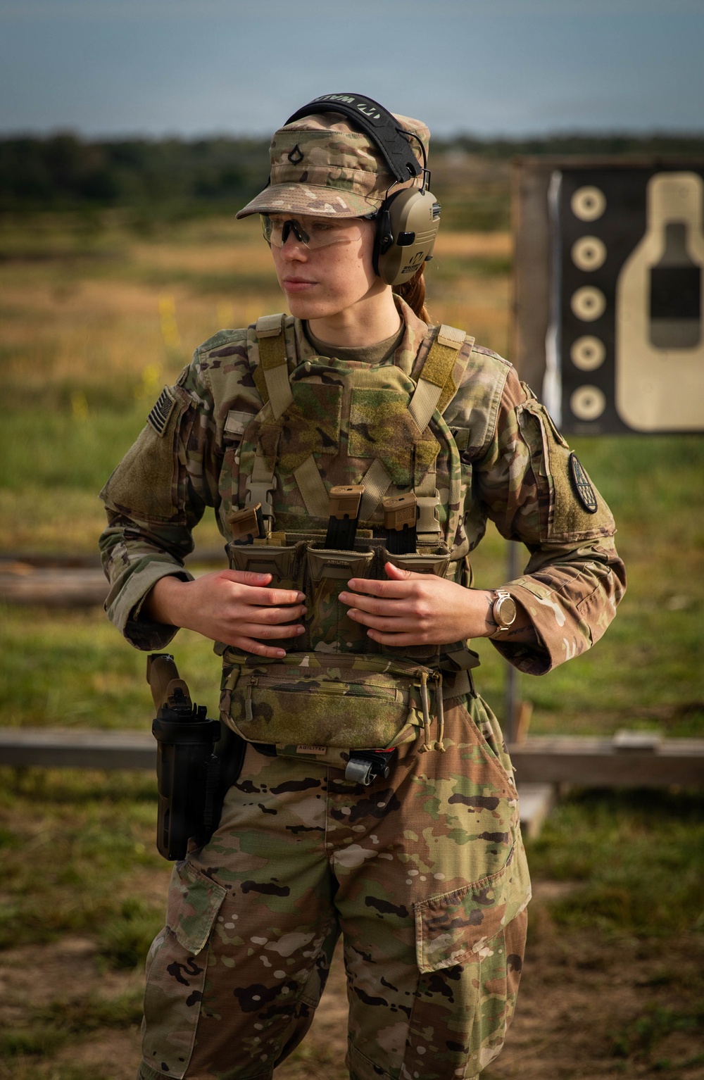 A soldier in U.S. Army OCP (Operational Camouflage Pattern) uniform stands at what appears to be a firing range. The individual is equipped with full tactical gear including a plate carrier, holstered pistol, and hearing/eye protection. A silhouette shooting target is visible in the background.