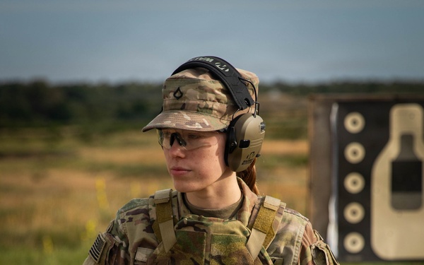 A soldier in U.S. Army OCP (Operational Camouflage Pattern) uniform stands at what appears to be a firing range. The individual is equipped with full tactical gear including a plate carrier, holstered pistol, and hearing/eye protection. A silhouette shooting target is visible in the background.