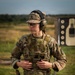 A soldier in U.S. Army OCP (Operational Camouflage Pattern) uniform stands at what appears to be a firing range. The individual is equipped with full tactical gear including a plate carrier, holstered pistol, and hearing/eye protection. A silhouette shooting target is visible in the background.
