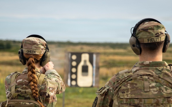 Two military personnel in MultiCam/OCP camouflage uniforms are at an outdoor firing range. The soldier on the left, identified by name tape as 'GHEEN,' is actively firing a pistol at a silhouette target downrange, while 'FIELITZ' on the right observes. Both are wearing hearing protection and tactical gear including plate carriers and backpacks.