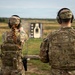 Two military personnel in MultiCam/OCP camouflage uniforms are at an outdoor firing range. The soldier on the left, identified by name tape as 'GHEEN,' is actively firing a pistol at a silhouette target downrange, while 'FIELITZ' on the right observes. Both are wearing hearing protection and tactical gear including plate carriers and backpacks.