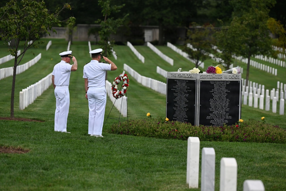 Two U.S. Navy personnel in white dress uniforms render a salute before the Pentagon September 11, 2001 memorial marker at Arlington National Cemetery. A floral wreath has been placed in front of the black granite memorial tablet, which bears the names of those killed at the Pentagon on 9/11. Rows of white headstones extend across the well-maintained cemetery grounds in the background.