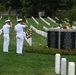 Two U.S. Navy personnel in white dress uniforms render a salute before the Pentagon September 11, 2001 memorial marker at Arlington National Cemetery. A floral wreath has been placed in front of the black granite memorial tablet, which bears the names of those killed at the Pentagon on 9/11. Rows of white headstones extend across the well-maintained cemetery grounds in the background.