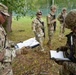 U.S. Army soldiers are engaged in what appears to be a field training exercise or operational briefing outdoors. Two soldiers in the foreground are reviewing documents on clipboards, while three additional soldiers stand in the background armed with rifles. All personnel are wearing OCP (Operational Camouflage Pattern) uniforms with U.S. flag patches.