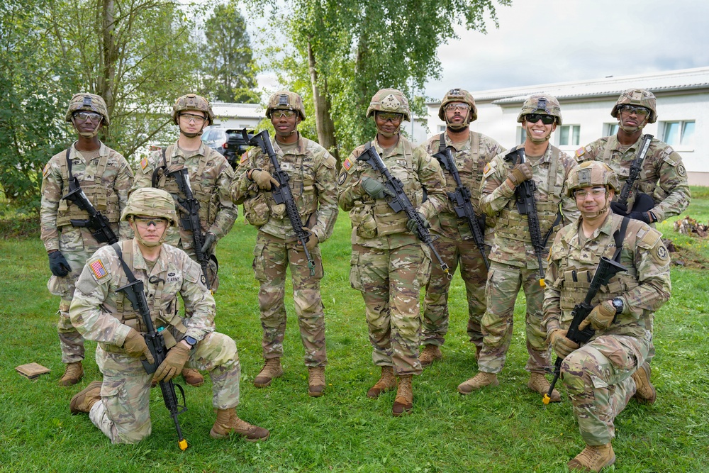 A group of approximately nine U.S. Army soldiers pose for a group photo outdoors on a grassy area. They are wearing MultiCam (OCP) camouflage uniforms, combat helmets, and protective eyewear, and are holding M4/M16-style rifles. Two soldiers are kneeling in the front row while the remaining soldiers stand behind them.