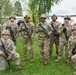 A group of approximately nine U.S. Army soldiers pose for a group photo outdoors on a grassy area. They are wearing MultiCam (OCP) camouflage uniforms, combat helmets, and protective eyewear, and are holding M4/M16-style rifles. Two soldiers are kneeling in the front row while the remaining soldiers stand behind them.