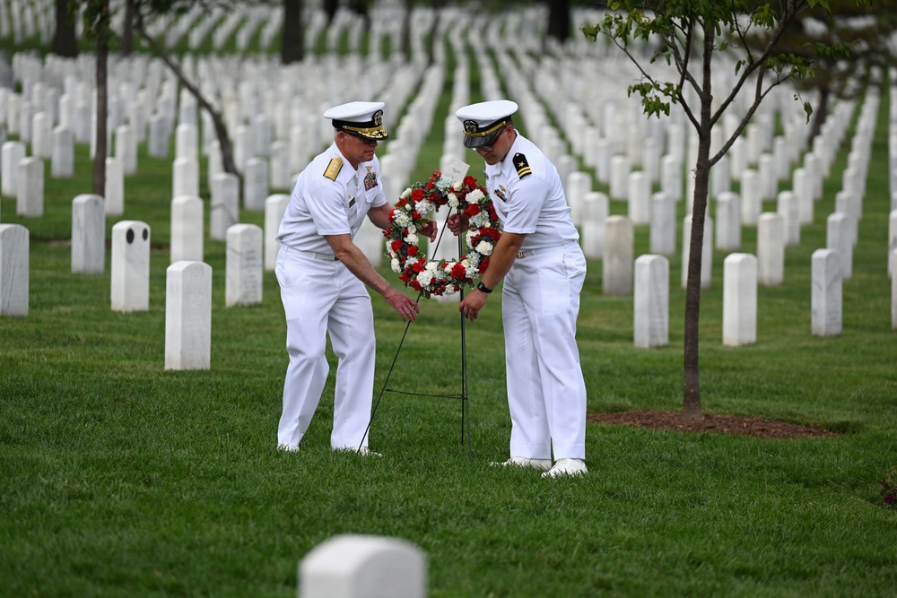 Two U.S. Navy officers in white dress uniforms are placing a red and white floral wreath on a stand among rows of white grave markers at what appears to be a military cemetery. The ceremony reflects a formal memorial tribute, likely occurring during a commemorative event such as Memorial Day. The setting features well-maintained green grass, uniform white headstones extending into the background, and young trees.