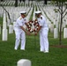 Two U.S. Navy officers in white dress uniforms are placing a red and white floral wreath on a stand among rows of white grave markers at what appears to be a military cemetery. The ceremony reflects a formal memorial tribute, likely occurring during a commemorative event such as Memorial Day. The setting features well-maintained green grass, uniform white headstones extending into the background, and young trees.