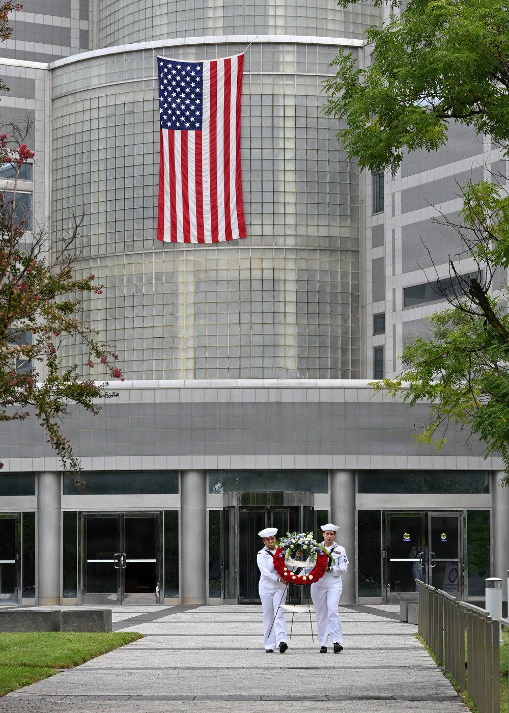 Two U.S. Navy sailors in dress white uniforms carry a floral wreath on a stand along a walkway toward a large modern building. A large American flag is prominently displayed vertically on the cylindrical facade of the building. The scene appears to be a formal memorial or commemorative ceremony.