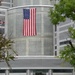 Two U.S. Navy sailors in dress white uniforms carry a floral wreath on a stand along a walkway toward a large modern building. A large American flag is prominently displayed vertically on the cylindrical facade of the building. The scene appears to be a formal memorial or commemorative ceremony.