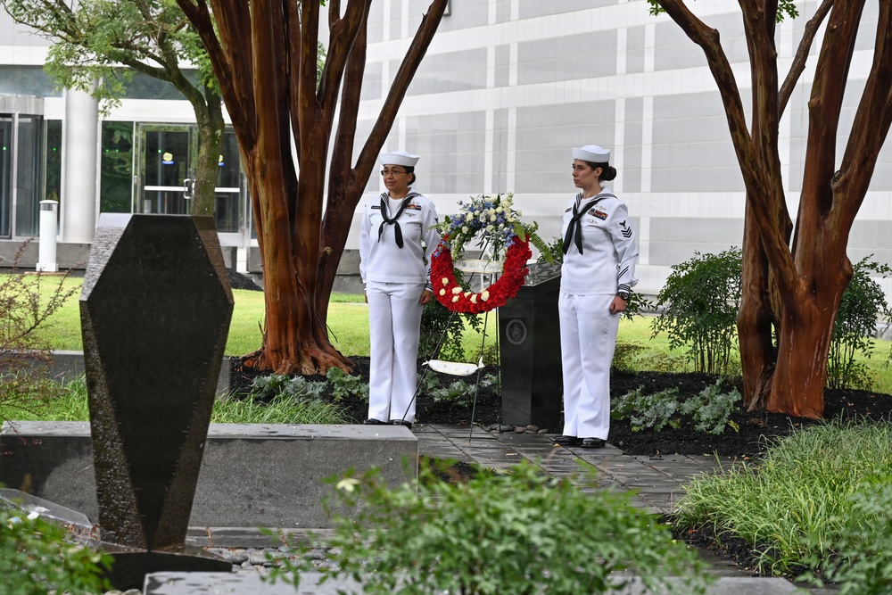Two U.S. Navy sailors in dress white uniforms stand at attention flanking a memorial wreath at an outdoor memorial site. The wreath features red and white flowers and is positioned in front of a dark stone monument. The setting appears to be a formal commemorative ceremony at what looks like a government or military installation.