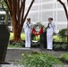 Two U.S. Navy sailors in dress white uniforms stand at attention flanking a memorial wreath at an outdoor memorial site. The wreath features red and white flowers and is positioned in front of a dark stone monument. The setting appears to be a formal commemorative ceremony at what looks like a government or military installation.