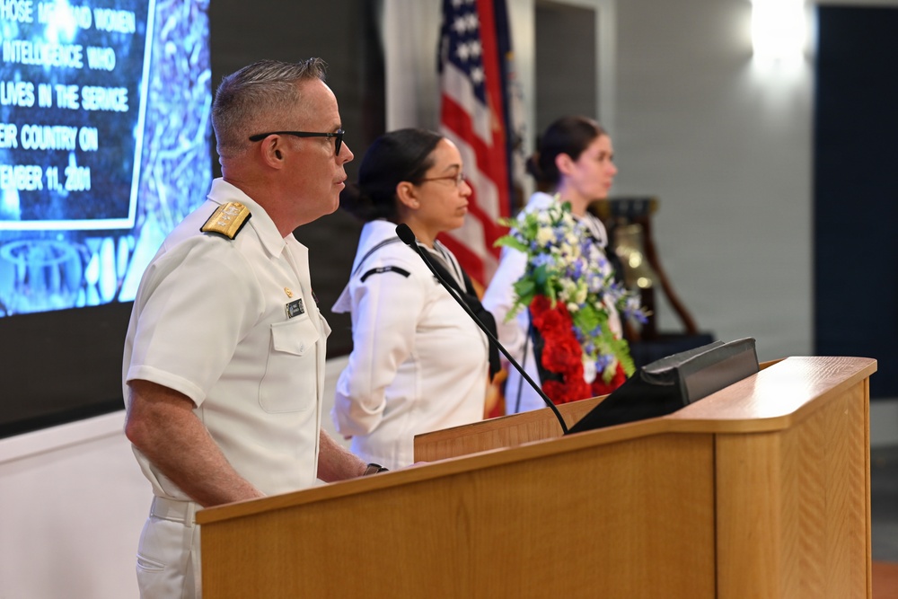 A military officer in white dress uniform stands at a wooden podium during what appears to be a September 11 memorial ceremony. Behind him, two individuals in white uniforms stand near a floral arrangement of red, white, and blue flowers, with an American flag visible in the background. A projection screen displays text referencing intelligence personnel who lost their lives on September 11, 2001.