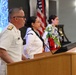 A military officer in white dress uniform stands at a wooden podium during what appears to be a September 11 memorial ceremony. Behind him, two individuals in white uniforms stand near a floral arrangement of red, white, and blue flowers, with an American flag visible in the background. A projection screen displays text referencing intelligence personnel who lost their lives on September 11, 2001.
