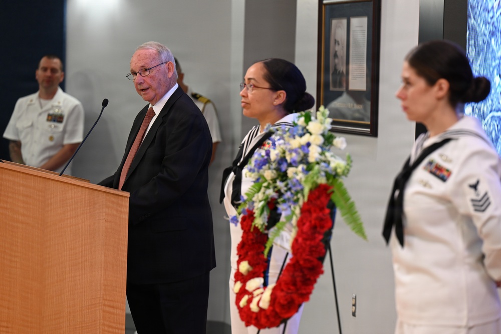 A formal military memorial ceremony is taking place indoors, likely at a naval installation or museum. An elderly civilian man in a dark suit is speaking at a podium while two female sailors in dress white uniforms stand at attention, one holding a large floral wreath composed of red, white, and blue flowers. Additional naval personnel in white uniforms are visible in the background.