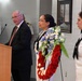 A formal military memorial ceremony is taking place indoors, likely at a naval installation or museum. An elderly civilian man in a dark suit is speaking at a podium while two female sailors in dress white uniforms stand at attention, one holding a large floral wreath composed of red, white, and blue flowers. Additional naval personnel in white uniforms are visible in the background.