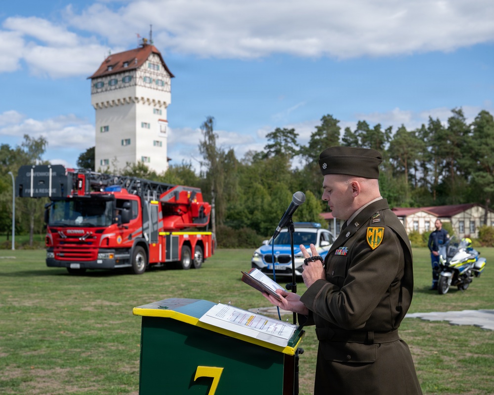A military officer in olive/green dress uniform stands at a podium reading from documents during what appears to be an official outdoor ceremony. The setting includes emergency vehicles — a red fire truck labeled 'Fire &amp; Rescue' and a police BMW motorcycle and vehicle — along with a distinctive white tower building with a half-timbered upper section in the background. The event appears to take place on a military or government installation, likely in Germany based on the architectural and vehicle styles.