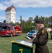 A military officer in olive/green dress uniform stands at a podium reading from documents during what appears to be an official outdoor ceremony. The setting includes emergency vehicles — a red fire truck labeled 'Fire &amp; Rescue' and a police BMW motorcycle and vehicle — along with a distinctive white tower building with a half-timbered upper section in the background. The event appears to take place on a military or government installation, likely in Germany based on the architectural and vehicle styles.