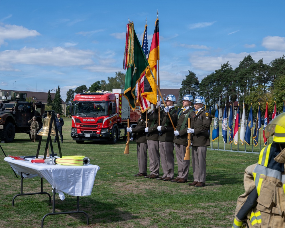 A formal military/civil service ceremony is taking place outdoors on a grassy field, featuring a color guard holding multiple flags including the American flag and German flag. A red Scania fire rescue truck labeled 'Fire Rescue Bavaria' is prominently displayed in the background, suggesting this is a fire department ceremonial event at a military installation in Bavaria, Germany. A table with firefighting equipment (hose, nozzle) and a ceremonial brass bell are positioned in the foreground.