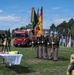 A formal military/civil service ceremony is taking place outdoors on a grassy field, featuring a color guard holding multiple flags including the American flag and German flag. A red Scania fire rescue truck labeled 'Fire Rescue Bavaria' is prominently displayed in the background, suggesting this is a fire department ceremonial event at a military installation in Bavaria, Germany. A table with firefighting equipment (hose, nozzle) and a ceremonial brass bell are positioned in the foreground.