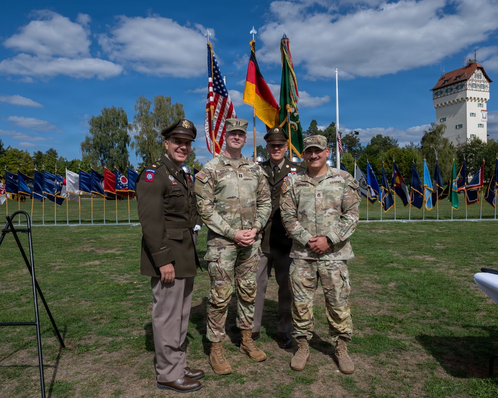Four U.S. Army personnel pose for a group photograph at an outdoor military ceremony. The setting includes a large display of unit guidons and national flags, including the U.S. and German flags, with a historic white tower building visible in the background. One soldier wears a vintage-style WWII-era Army dress uniform with an 82nd Airborne patch, while the others wear modern Army Combat Uniforms (ACU).