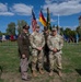 Four U.S. Army personnel pose for a group photograph at an outdoor military ceremony. The setting includes a large display of unit guidons and national flags, including the U.S. and German flags, with a historic white tower building visible in the background. One soldier wears a vintage-style WWII-era Army dress uniform with an 82nd Airborne patch, while the others wear modern Army Combat Uniforms (ACU).