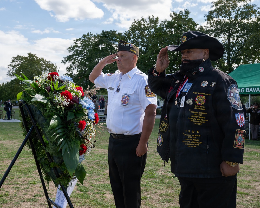 Two veterans are saluting during a wreath-laying ceremony at an outdoor memorial event. The man on the left wears a white uniform shirt with a cap reading 'EUROPE', while the man on the right wears a black jacket adorned with numerous patches and medals, including VFW insignia, and a wide-brimmed black hat. A floral wreath featuring red, white, and blue flowers is displayed on a stand in the foreground.