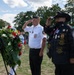 Two veterans are saluting during a wreath-laying ceremony at an outdoor memorial event. The man on the left wears a white uniform shirt with a cap reading 'EUROPE', while the man on the right wears a black jacket adorned with numerous patches and medals, including VFW insignia, and a wide-brimmed black hat. A floral wreath featuring red, white, and blue flowers is displayed on a stand in the foreground.
