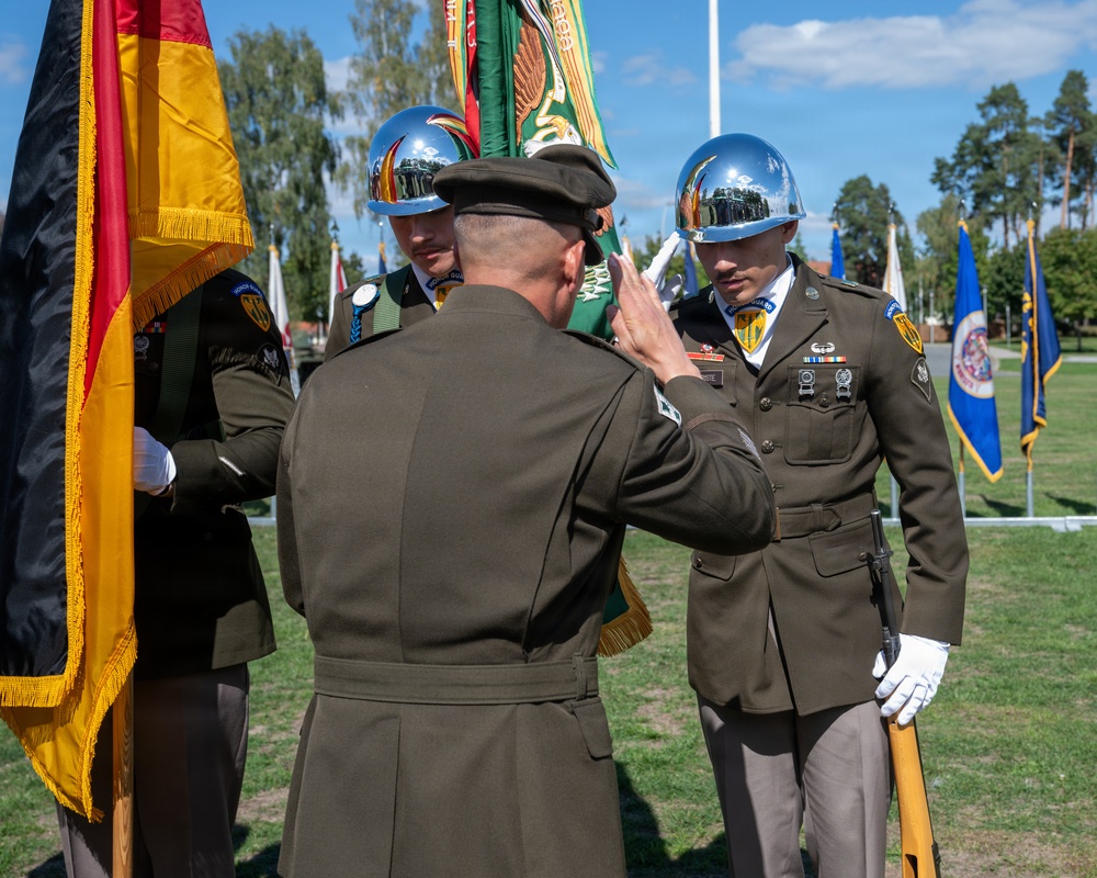A military ceremony is taking place outdoors on a well-maintained parade ground. A senior officer with his back to the camera is receiving a salute from a soldier in dress uniform wearing a chrome ceremonial helmet, while color guard members hold flags including the German national flag. The setting appears to be a formal change of command or honors ceremony at a military installation.