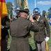 A military ceremony is taking place outdoors on a well-maintained parade ground. A senior officer with his back to the camera is receiving a salute from a soldier in dress uniform wearing a chrome ceremonial helmet, while color guard members hold flags including the German national flag. The setting appears to be a formal change of command or honors ceremony at a military installation.