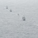 An aerial photograph shows five naval vessels sailing in a loose diagonal formation across open ocean. The ships appear to be gray-painted military vessels of varying sizes, likely conducting a joint naval exercise or patrol operation. The image is taken from altitude under overcast conditions, giving the scene a muted, hazy appearance.