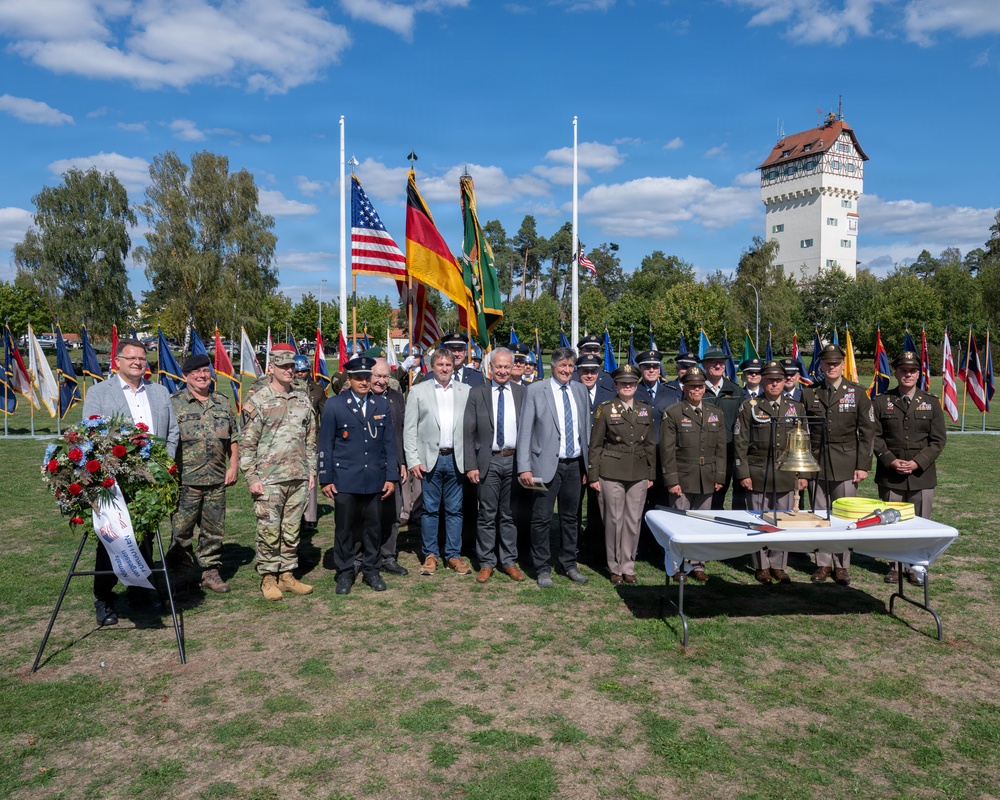 A formal outdoor ceremony featuring a mixed group of military personnel in various uniforms and civilians posing together on a grass field. Both American and German flags are prominently displayed alongside numerous military unit colors and guidons. The setting appears to be a military installation in Germany, identifiable by the distinctive white tower with a red-tiled roof in the background.