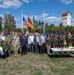 A formal outdoor ceremony featuring a mixed group of military personnel in various uniforms and civilians posing together on a grass field. Both American and German flags are prominently displayed alongside numerous military unit colors and guidons. The setting appears to be a military installation in Germany, identifiable by the distinctive white tower with a red-tiled roof in the background.