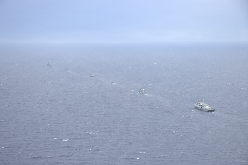 An aerial photograph shows a naval formation of approximately 7-8 warships sailing in a single-file line column across open ocean. The vessels appear to be gray-painted military ships of varying sizes, photographed from an elevated vantage point, likely from an aircraft. Hazy atmospheric conditions reduce visibility, particularly for the more distant vessels in the formation.