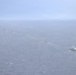 An aerial photograph shows a naval formation of approximately 7-8 warships sailing in a single-file line column across open ocean. The vessels appear to be gray-painted military ships of varying sizes, photographed from an elevated vantage point, likely from an aircraft. Hazy atmospheric conditions reduce visibility, particularly for the more distant vessels in the formation.