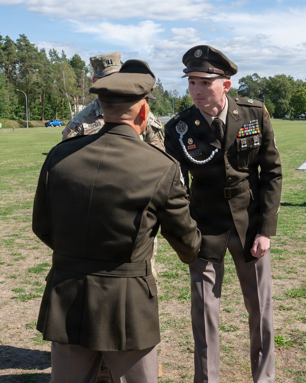 A U.S. Army military ceremony taking place outdoors on a grass field. A soldier in Army Green Service Uniform (Class A) with multiple medals and a distinctive unit insignia is facing a senior officer whose back is to the camera, suggesting a formal presentation or award ceremony. A third soldier in Army Combat Uniform (ACU) with the name tape 'MAPLES' is visible in the background.