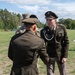 A U.S. Army military ceremony taking place outdoors on a grass field. A soldier in Army Green Service Uniform (Class A) with multiple medals and a distinctive unit insignia is facing a senior officer whose back is to the camera, suggesting a formal presentation or award ceremony. A third soldier in Army Combat Uniform (ACU) with the name tape 'MAPLES' is visible in the background.