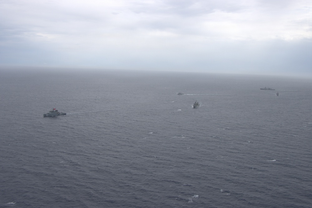 An aerial photograph showing a formation of multiple naval vessels underway on an overcast, grey sea. At least five ships are visible spread across the frame, suggesting a multi-ship naval exercise or transit operation. Visibility is reduced due to hazy, overcast weather conditions.