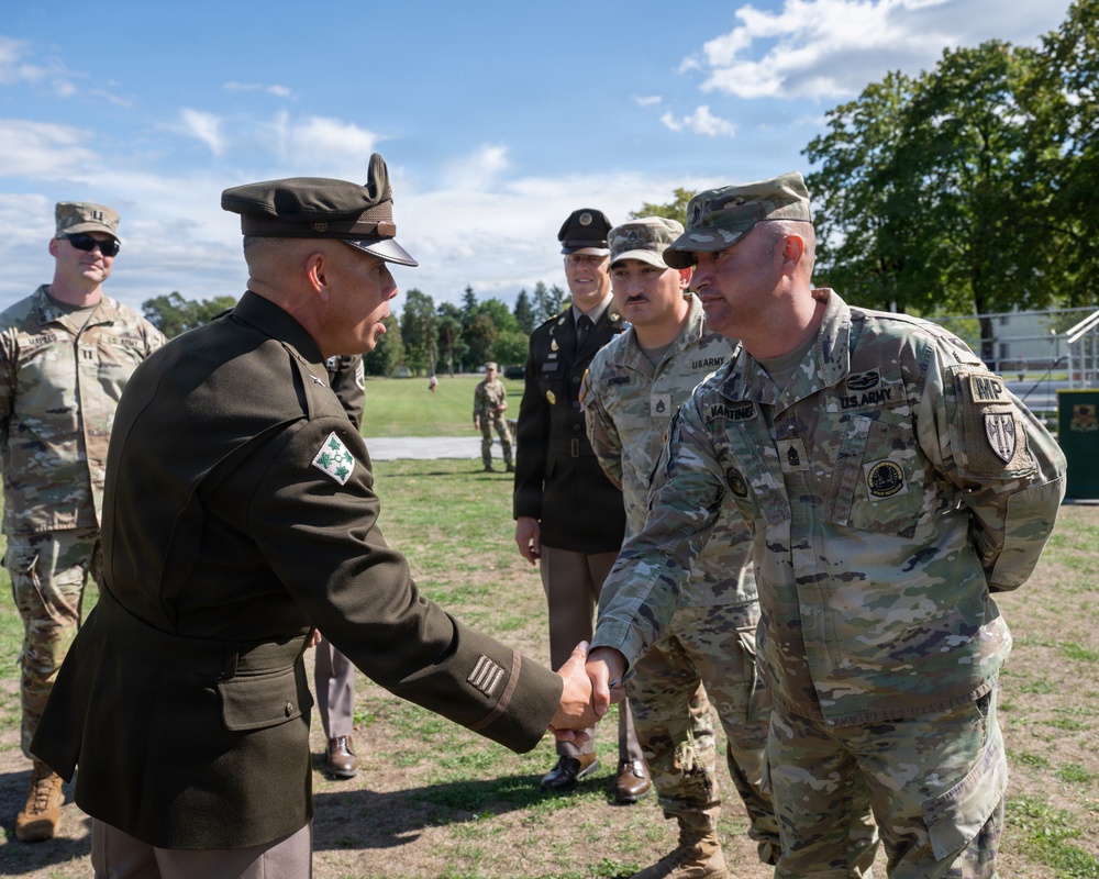 A military ceremony or formal meeting is taking place outdoors on a grassy field. An officer in an olive/dark green dress uniform is shaking hands with a U.S. Army soldier in OCP (Operational Camouflage Pattern) combat uniform. Several other military personnel in both dress and combat uniforms are present in the background observing the exchange.