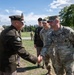A military ceremony or formal meeting is taking place outdoors on a grassy field. An officer in an olive/dark green dress uniform is shaking hands with a U.S. Army soldier in OCP (Operational Camouflage Pattern) combat uniform. Several other military personnel in both dress and combat uniforms are present in the background observing the exchange.