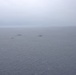 An aerial or elevated photograph showing a formation of approximately six naval vessels spread across an open ocean under overcast, hazy conditions. The ships appear to be operating in a loose formation, possibly during a naval exercise or patrol. Weather conditions are poor with low visibility and a grey, cloud-covered sky.